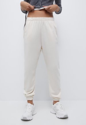 Osoba nosząca białe joggery z elastyczną talią i białe trampki, poprawiająca szary krótki kardigan, stojąca na tle jednolitego jasnego tła.