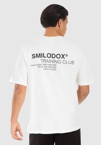 Hvid bomulds T-shirt med sort trykt tekst på ryggen: "SMILODOX® TRAINING CLUB" sammen med yderligere motiverende sætninger. Korte ærmer.