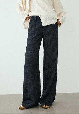 Personne portant un pantalon large en denim foncé avec des sandales marron à bout ouvert et une veste oversize beige clair, debout sur un sol gris.