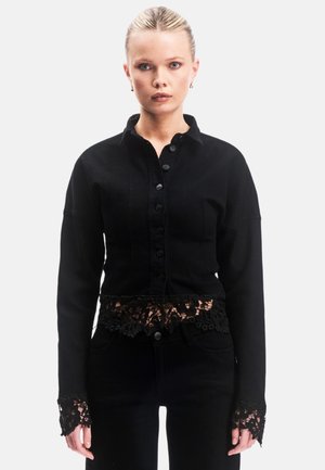 Frau mit blondem Haar in einem schwarzen, geknöpften Hemd mit Spitzen-Details am Saum und den Manschetten, trägt schwarze Hose vor weißem Hintergrund.