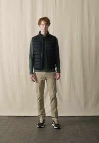 Schwarze Steppweste über einem dunkelgrünen Langarmshirt, kombiniert mit hellbeigen Hosen und schwarzen Sneakers, steht vor einem neutralen Hintergrund.