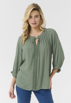 Blouse verte en tissu texturé, dotée d'un col rond avec un lien, des manches 3/4 et des coutures décoratives à l'avant.