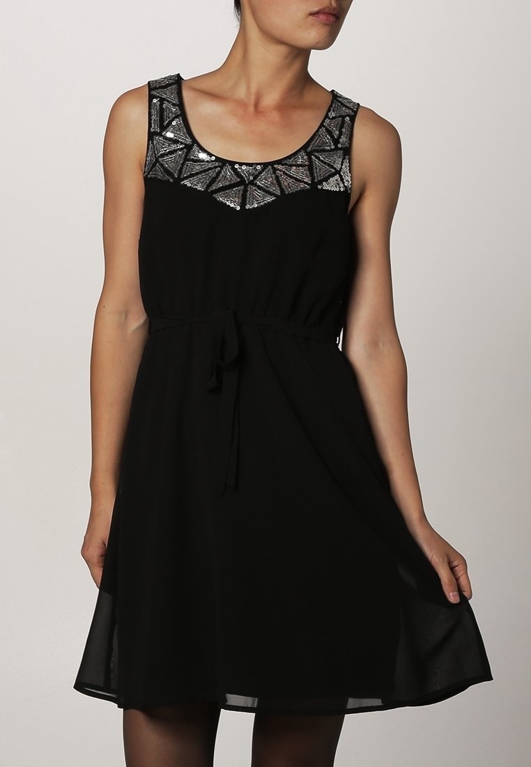 Femme portant une robe noire sans manches avec un détail géométrique en sequins argentés au niveau du décolleté, tenant l'ourlet d'une main.