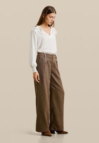 Blouse blanche à manches longues avec un col en V et des poignets smockés, associée à un pantalon large marron taille haute avec un motif subtil.