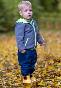 Kind in einer grauen Jacke mit einer grünen Kapuze, blauen wasserdichten Hosen und gelben Gummistiefeln mit braunen Akzenten, steht auf gefallenen Blättern.