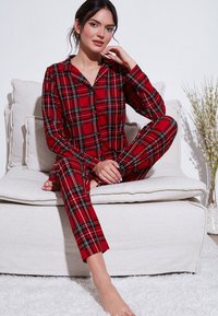 Punainen ruudullinen puuvillapukusarja, jossa on napitettava paita ja pitkät housut mustilla ja vihreillä yksityiskohdilla, neutraalin taustan edessä.