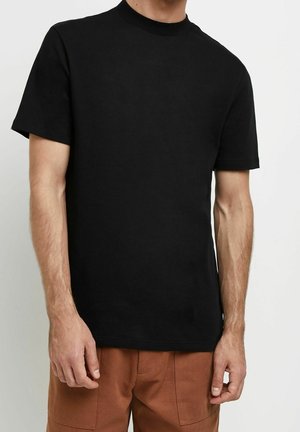 Mann in einem schlichten schwarzen T-Shirt mit kurzen Ärmeln und braunen Hosen, der vor einem weißen Hintergrund steht.