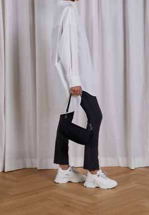 Witte oversized blouse, zwarte broek met fijne strepen en dikke witte sneakers. Zwarte handtas met riem in de hand. Houten vloer en neutrale achtergrond.