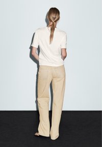 Una camicia bianca a maniche corte abbinata a pantaloni dritti beige chiaro, con tasche posteriori e una vestibilità rilassata, su uno sfondo neutro.