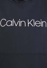 Tmavě modrá mikina s kapucí vyrobená z měkkého materiálu. Obsahuje velké bílé logo "Calvin Klein" a klokaní kapsu na přední straně.