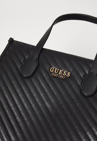Sac cabas en cuir noir avec une texture côtelée. Présente un logo en métal doré "GUESS EST 1981" à l'avant. Comprend deux poignées supérieures.