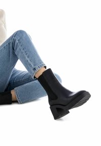 Botín negro de tacón con paneles laterales elásticos, suela texturada, puntera redonda y tacón grueso, combinado con jeans de lavado claro.