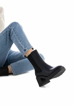 Botín negro de tacón con paneles laterales elásticos, suela texturada, puntera redonda y tacón grueso, combinado con jeans de lavado claro.