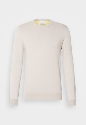 Jasno beżowy sweter dzianinowy z długimi rękawami, okrągłym dekoltem i subtelnymi ściągaczami. Posiada małe złote logo na klatce piersiowej.