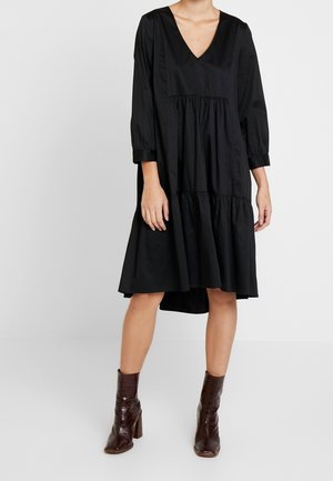 Femme portant une robe noire ample à col en V avec des manches trois-quarts et des bottines en cuir marron, debout sur un sol blanc uni.