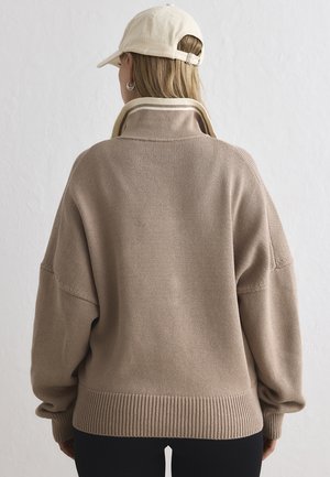 Personne portant un pull en tricot beige à col montant et une casquette de baseball beige, vue de dos sur un fond uni.