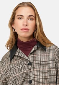 Geruit overhemdjack met zwarte leren kraag, met een beige en roze ruitpatroon en zwarte knopen. Coltrui in bordeauxrood eronder.