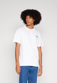 Bílý bavlněný tričko s krátkým rukávem, kulatým výstřihem a malým černým logem "TOMMY JEANS" na levé hrudi, nošeno s modrými džínami.