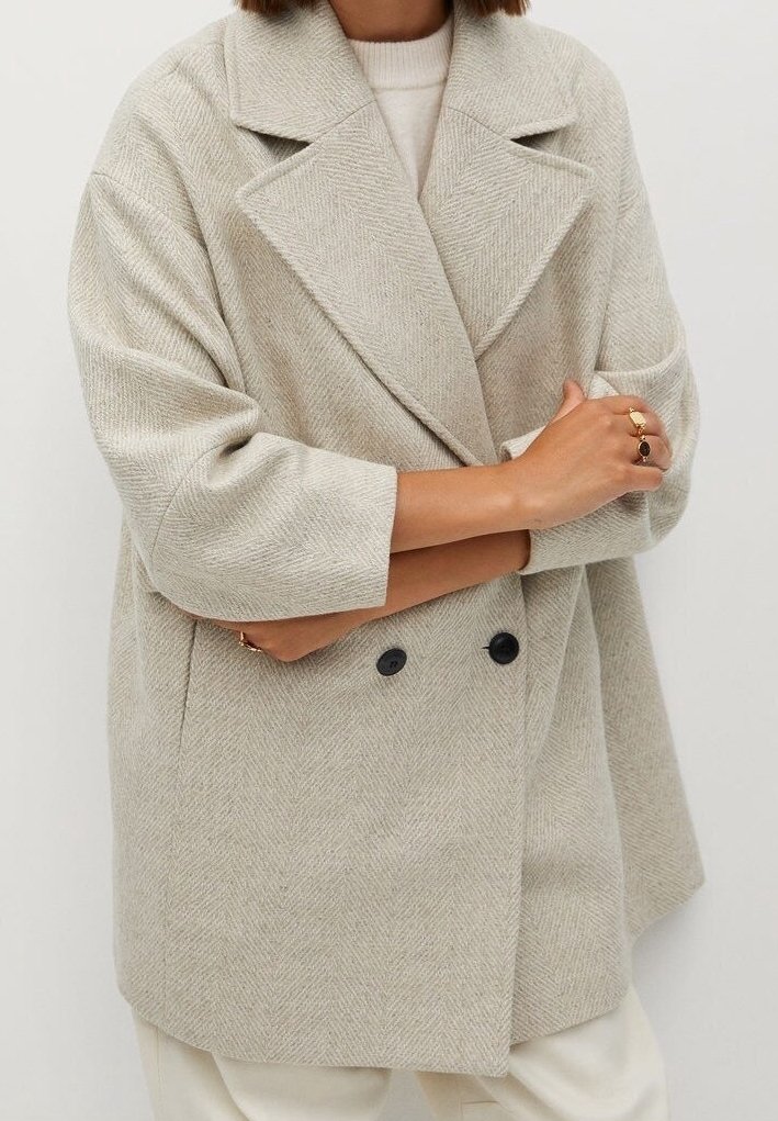 Femme portant un manteau en laine beige clair surdimensionné à double boutonnage avec de larges revers, manches retroussées, et deux boutons noirs, les bras croisés.