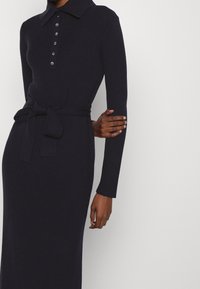 Stickad långärmad klänning i mörk marinblå med krage, knappstängning fram och ett tygbälte i midjan.