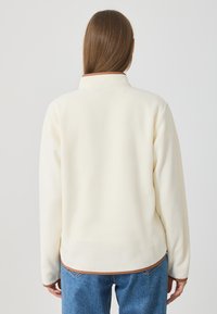 Jachetă din fleece crem, cu guler înalt și margine maro, având o textură netedă și o croială lejeră, purtată peste blugi albaștri.