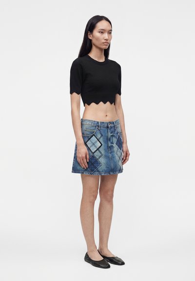 Γυναίκα στέκεται φορώντας μαύρο κοντομάνικο crop top με κυματιστό τελείωμα, μπλε patchwork τζιν μίνι φούστα και μαύρα φλατ παπούτσια σε απλό φόντο.