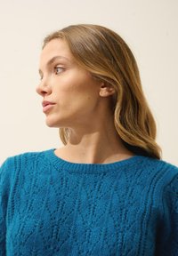 Teal stickad tröja med en texturerad diamantsstruktur, rund halsringning och ribbade ärmslut. Mjuk tyg med en lätt känsla.