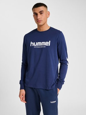 Młody mężczyzna ubrany w granatową bluzę hummel z długim rękawem i pasujące granatowe spodnie dresowe z logo na piersi i udzie stoi na jednolitym tle.