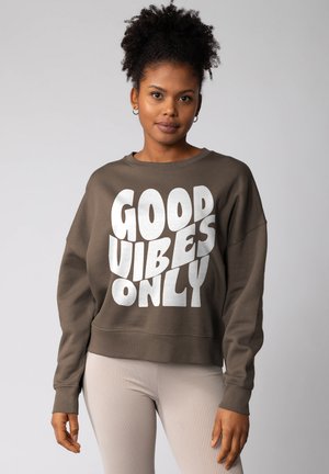 Junge Frau mit lockigem Haar trägt einen braunen Sweatshirt mit der Aufschrift "Good Vibes Only" und helle, gerippte Leggings, während sie vor einem schlichten grauen Hintergrund steht.