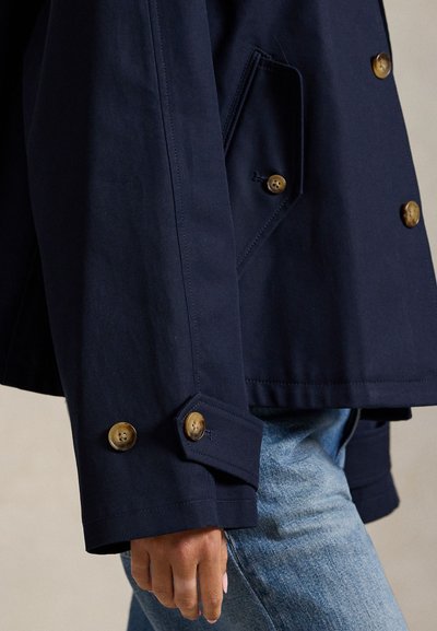 Μπλε-ναυτικό βαμβακερό μπουφάν με διπλό πέτο, κουμπιά χρυσού χρώματος και λοξά μανίκια. Λεπτομέρεια τσέπης ορατή στο πλάι.