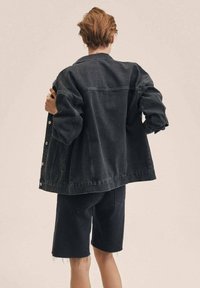 Persona con capelli corti che indossa una giacca di denim nera oversize e pantaloncini sfilacciati abbinati, voltata di spalle su uno sfondo semplice.