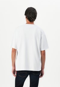 Man with dark hair wearing a plain white short-sleeve shirt and dark jeans, standing facing a plain white background.