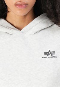Žena v svetlosivej mikine s logom Alpha Industries na hrudi, s uzatvorenými perami a kučeravými tmavými vlasmi viditeľnými z boku.