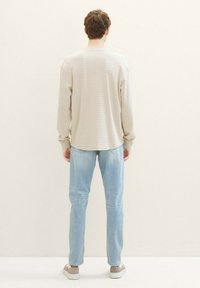 Beige Langarmshirt mit vertikalen Streifen, Rundhals, kombiniert mit hellblauen Jeans und grauen Sneakern, von hinten betrachtet.