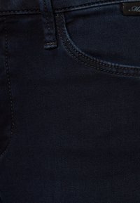 Dark blue denim fabric of jeans, featuring a smooth texture, standard stitching, and a small brand label at the waistband.