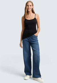 Czarna koszulka na cienkich ramiączkach, zestawiona z szerokimi niebieskimi dżinsami z szerokimi nogawkami. Do stroju dodano białe trampki. Tło jest jasnoszare.