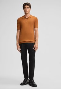 Polo a maniche corte di colore arancione bruciato, in tessuto a maglia liscia, con colletto e patta con tre bottoni, abbinato a pantaloni neri affusolati e scarpe.