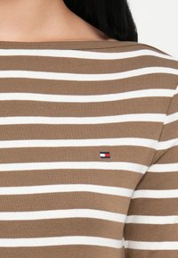 Καφέ και λευκή ριγάτη μπλούζα με μακριά μανίκια, ribbed λαιμό με μικρό κεντημένο λογότυπο στην αριστερή πλευρά. Μαλακό ύφασμα από μίγμα βαμβακιού.