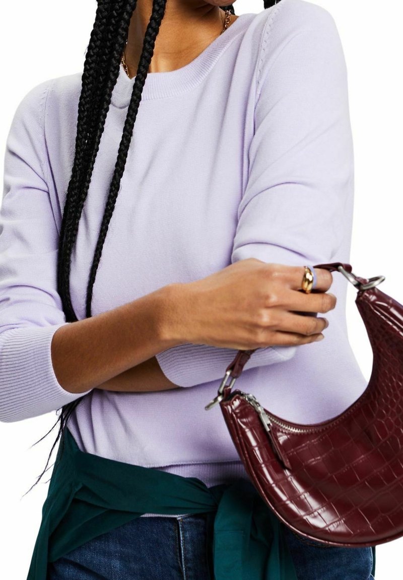 Femme aux longues tresses portant un pull lavande, tenant un sac à main bordeaux à motif croco et une veste vert foncé nouée à la taille.