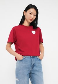 Tânără cu păr drept, negru, purtând un tricou roșu cu o patch alb în formă de inimă și blugi deschiși, rupti, cu mâinile în buzunare.