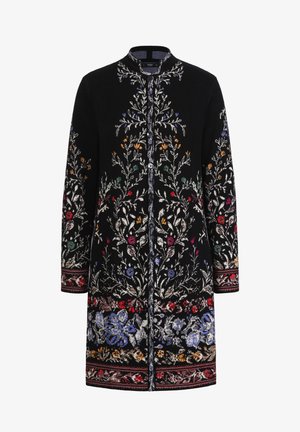 Melnā mētelī ar sarežģītu ziedu izšuvumu sarkanā, zilā un zaļā krāsā. Ar apaļu apkakli un pogu aizdari, kā arī ar rakstainām apakšmalas akcentiem.