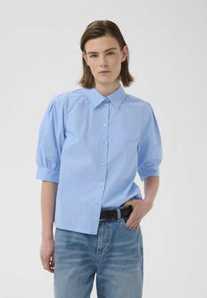 Femme aux cheveux courts et bruns portant une chemise bleu clair à manches bouffantes boutonnée et un jean bleu délavé avec une ceinture noire, sur un fond uni.