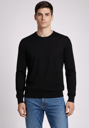 Jonge man draagt een zwarte trui met lange mouwen en een blauwe spijkerbroek, staand tegen een effen lichte achtergrond.