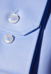 Lichtblauwe stoffen met een fijne ribbeltextuur, voorzien van twee doorzichtige knopen en scherpe kraagranden.