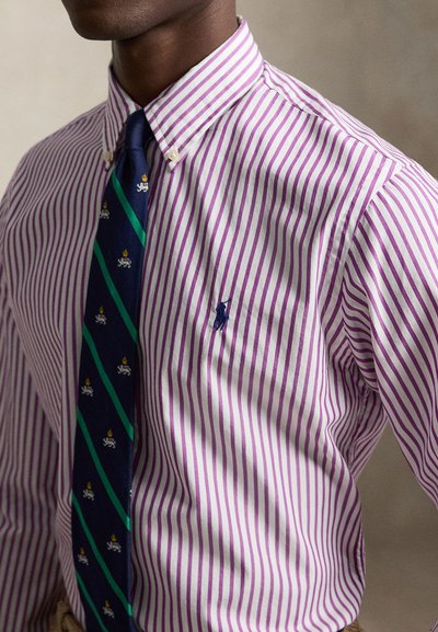 Homme portant une chemise à rayures violettes et blanches à boutons, avec une cravate bleu marine ornée de rayures vertes et de petits motifs blancs de lion et de couronne.