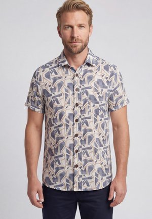 Blonder Mann trägt ein kurzärmeliges Hemd mit abstrakten blauen und beigen geometrischen Mustern, steht vor einem einfarbigen Hintergrund.