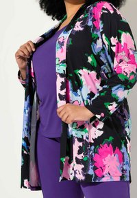 Cardigan con estampado floral en negro, con acentos en rosa, azul y verde. Combinado con una blusa púrpura y pantalones, hechos de una tela suave.