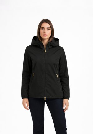 Donna che indossa una giacca nera con cappuccio e dettagli dorati sulla cerniera, in piedi con le braccia rilassate contro uno sfondo bianco uniforme.