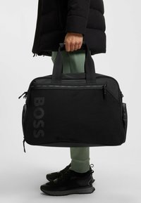 Borsa duffel nera con tessuto testurizzato, apertura con zip, manici doppi e logo "BOSS" in una tonalità più scura; progettata per durabilità e funzionalità.