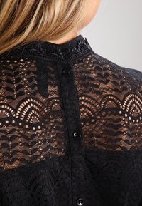 Sort blonder-top med indviklede mønstre, gennemsigtig tekstur og dekorativ knaplukning bagpå, med høj halsdesign.